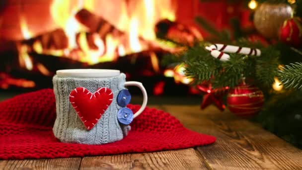 Šálek s horkým nápojem v modrém pleteném obalu s červeným srdcem na dřevěném stole na pozadí hořícího krbu, červeným kostkovaným a vánočním stromečkem. — Stock video