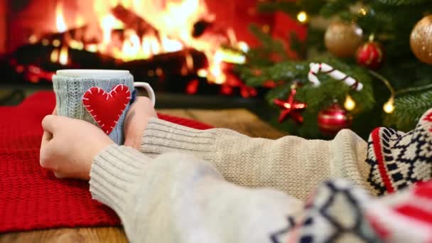 Womans hands in a warm sweater holds a cup with a hot drink in a blue knitted cover with a red heart against the background of a burning fireplace and a Christmas tree. — Stock Video