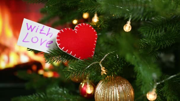 A red felt heart and a note with the text With Love on the background of a Christmas tree and a burning fireplace. — Vídeo de Stock