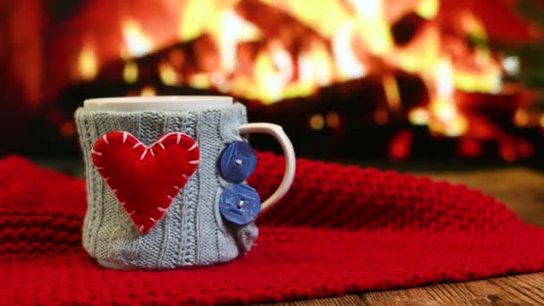 Šálek s horkým nápojem v modrém pleteném obalu s červeným srdcem na dřevěném stole na pozadí hořícího krbu a červeným kostkovaným — Stock video