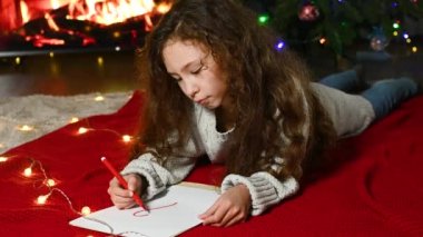 child girl with curly hair lies on a red knitted blanket against the background of a burning fireplace and draws a red heart on a white sheet of paper.
