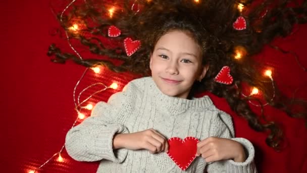 Baby girl with a garland and red hearts in curly hair smiles on a red background and holds a heart in her hands. — Stockvideo