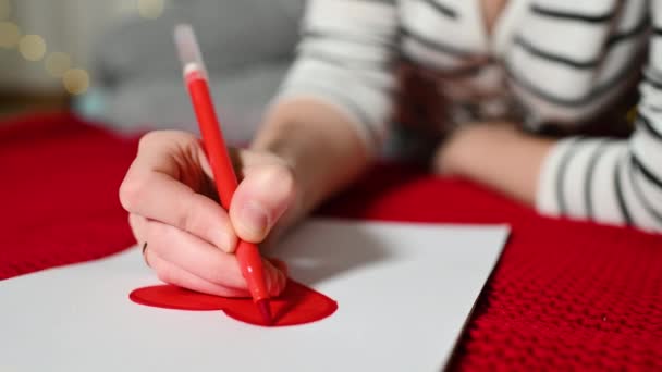 Jeune femme en pull rayé dessine un cœur rouge sur une feuille blanche de carte de Saint-Valentin et se trouve sur un plaid rouge. Saint Valentin — Video