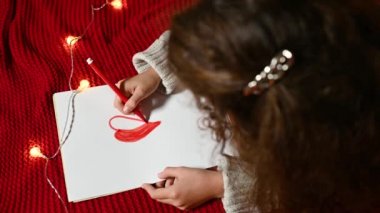Kıvırcık saçlı küçük bir kız sevgililer günü için beyaz bir kağıt parçasına kalp çiziyor kırmızı örgü bir ekose üzerinde yatıyor.