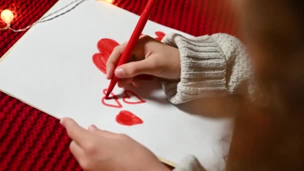 Malá holčička s kudrnatými vlasy nakreslí srdce na bílém kousku papíru na Valentýna ležícím na červeném pleteném kostkovaném. — Stock video