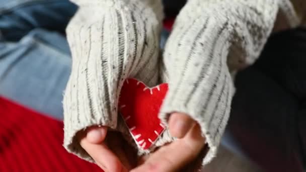 Close-up of the palms half-covered by the sleeves of a knitted sweater holding a small red heart in their hands. — Vídeo de stock