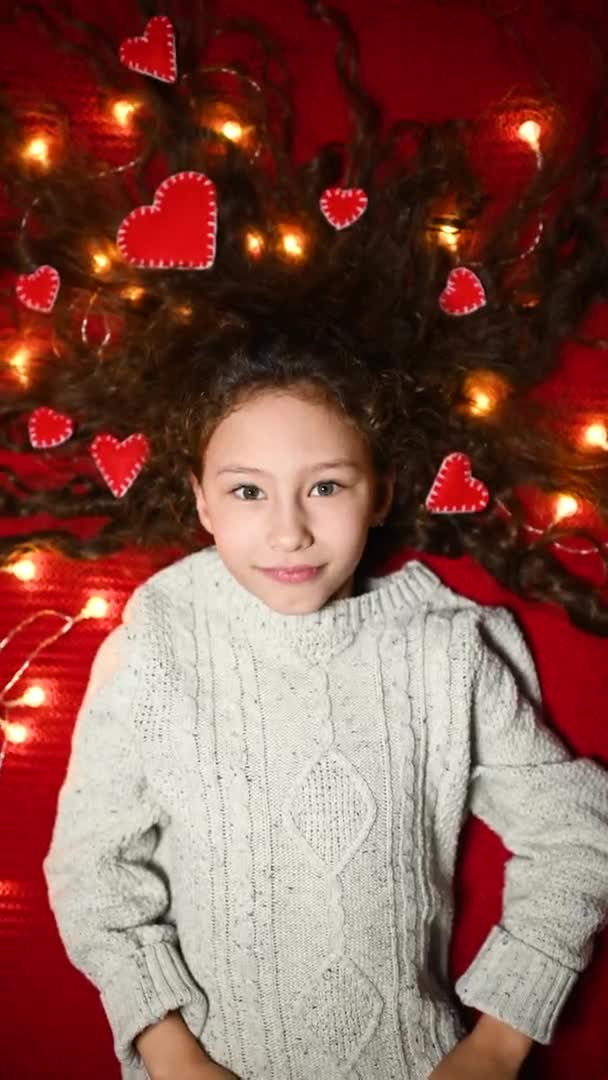Baby girl with a garland and red hearts in curly hair smiles on a red background and holds a heart in her hands — Vídeos de Stock