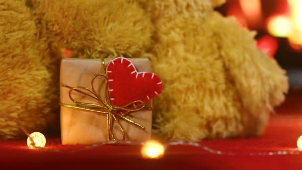 Пара плюшевих ведмедиків сидить обіймаючись на задньому плані каміну і тримаючи в лапах коробку з подарунком і червоним серцем. День Святого Валентина — стокове відео