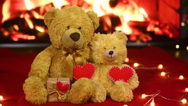 Pair of teddy bears are sitting hugging against the background of a red knitted plaid and a garland of light bulbs and holding two red hearts in their paws - a symbol of love. — стоковое видео