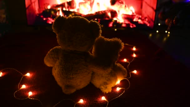 Two cute teddy bears hugging each other sitting on a knitted red plaid against the background of a garland of light bulbs and a burning fireplace — Vídeo de Stock