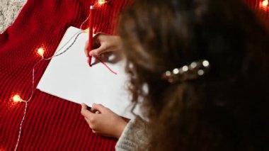 Kıvırcık saçlı küçük bir kız sevgililer günü için beyaz bir kağıt parçasına kalp çiziyor kırmızı örgü bir ekose üzerinde yatıyor.