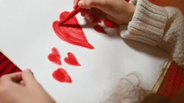 Kıvırcık saçlı küçük bir kız sevgililer günü için beyaz bir kağıt parçasına kalp çiziyor kırmızı örgü bir ekose üzerinde yatıyor.