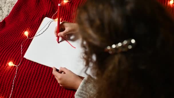 Una niña pequeña con el pelo rizado dibuja un corazón en un pedazo blanco de papel para el Día de San Valentín acostado en una tela a cuadros de punto rojo. — Vídeos de Stock