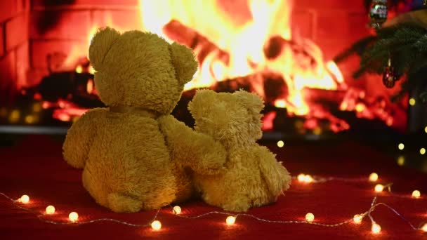 Two cute teddy bears hugging each other sitting on a knitted red plaid against the background of a garland of light bulbs and a burning fireplace — Αρχείο Βίντεο