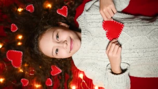 Liten flicka med en krans och röda hjärtan i lockigt hår ler på en röd bakgrund och håller ett hjärta i händerna — Stockvideo