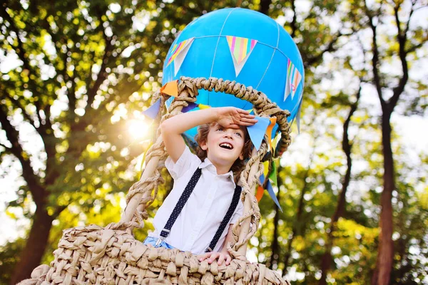 Мальчик с вьющимися волосами в корзине синего шарика улыбается и смотрит вдаль на фоне зелени и солнца. — стоковое фото