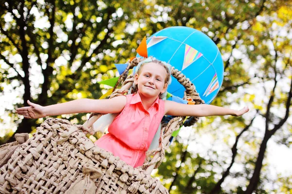 Liten flicka i rosa kläder leenden sitter i en korg med en blå ballong mot en bakgrund av solljus och grönska. — Stockfoto