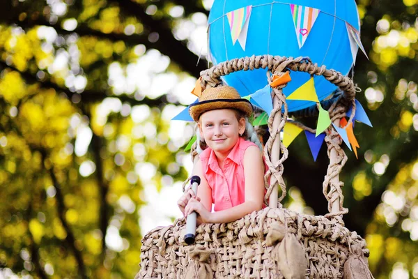 Liten flicka i rosa kläder med en halmhatt och ett teleskop i händerna ler i korgen på en blå ballong. — Stockfoto