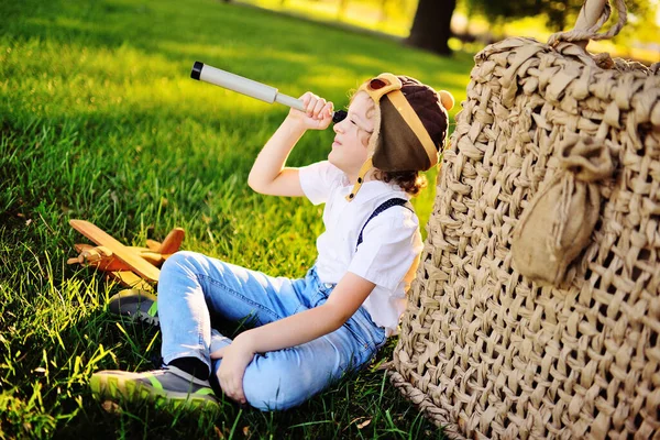 Маленький мальчик в белой рубашке с подтяжками и в шлеме пилота в очках улыбается, сидя на траве, опираясь на корзину из воздушного шара на фоне деревьев — стоковое фото