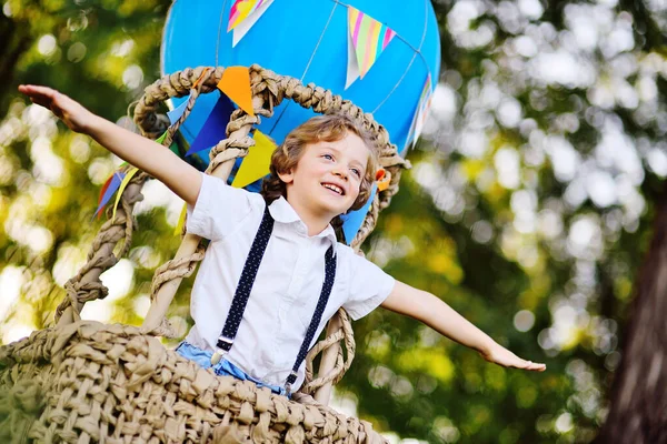 Kleine jongen met krullend haar in een mand van een blauwe ballon glimlacht en kijkt in de verte tegen de achtergrond van groen en de zon. — Stockfoto