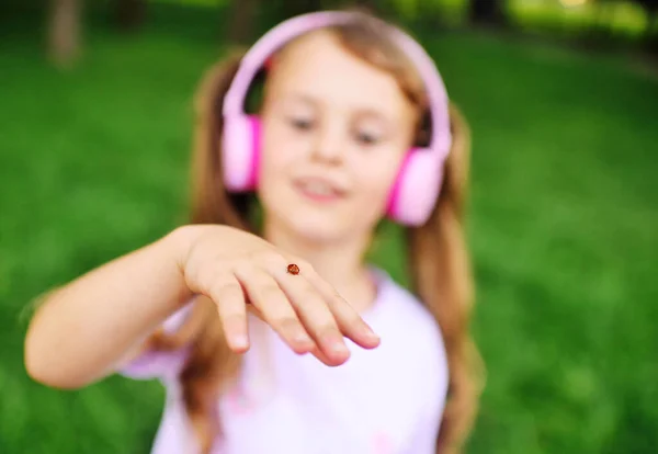 Uma menina em fones de ouvido rosa olha para uma joaninha em sua mão. — Fotografia de Stock