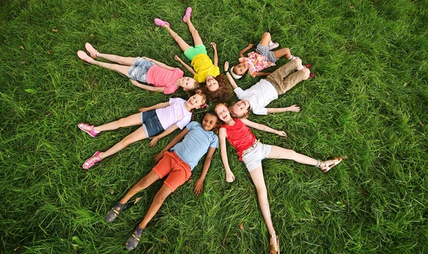 Farklı ırktan ve milletten çocuklar çimenlerin üzerinde renkli giysiler içinde uzanır ve gülümserler.. — Stok fotoğraf