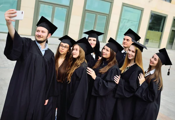 Groep van afgestudeerde studenten van meesters in zwarte gewaden en vierkante hoeden selfies nemen op een smartphone camera. — Stockfoto