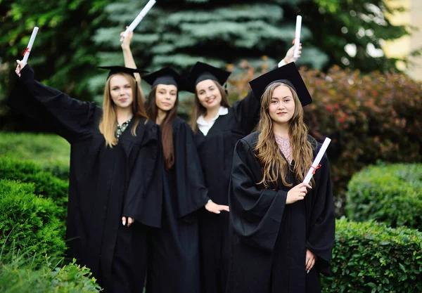 Bella giovane ragazza è una studentessa laureata in una veste nera sorridente in possesso di un diploma nelle sue mani sullo sfondo di un gruppo di laureati. — Foto Stock