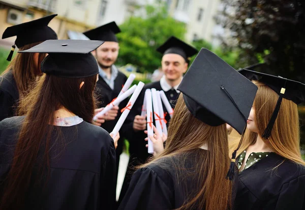 Groep van universitaire afgestudeerden in student gewaden of mantel en vierkante hoeden bij de diploma-uitreiking houden college diploma diploma 's in hun handen en verheugen zich. — Stockfoto