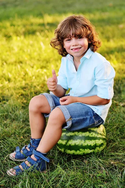 Мальчик с вьющимися волосами сидит на большом арбузе и улыбается в парке в солнечный день — стоковое фото
