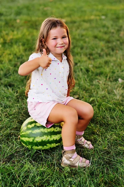 Маленькая забавная девочка с рыжими волосами, опирающаяся на огромный арбуз в парке на траве в летний день — стоковое фото