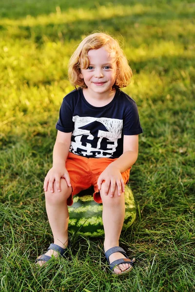 Niño con el pelo rizado se sienta en una gran sandía y sonríe en el parque en un día soleado — Foto de Stock