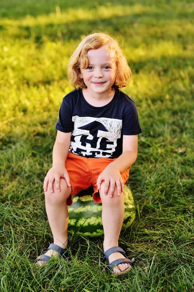 Baby boy with curly hair sits on a big watermelon and smiles in the Park on a Sunny day — Stock Photo, Image