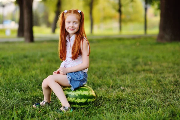 Рыжая девушка с арбузными гримасами и улыбками, сидящая на траве в парке — стоковое фото