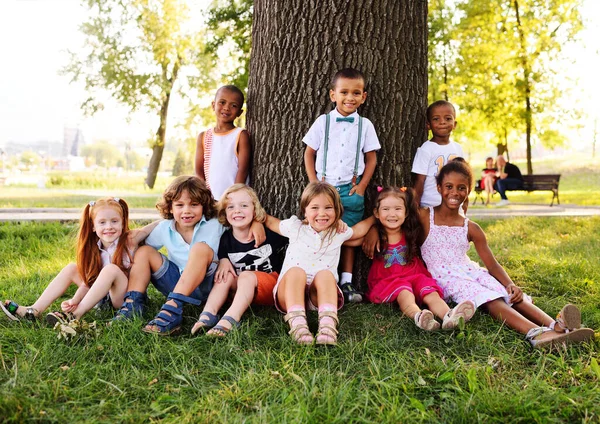 Um grupo de crianças pequenas em roupas coloridas abraçando sentado na grama sob uma árvore em um parque rindo e sorrindo . — Fotografia de Stock