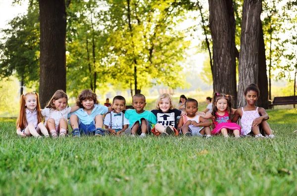 Muchos niños pequeños de diferentes razas juegan juntos en el parque en la hierba fresca verde. — Foto de Stock
