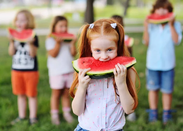 Рыжая маленькая девочка жадно ест сочный спелый арбуз на фоне своих друзей и Парк в солнечный летний день — стоковое фото