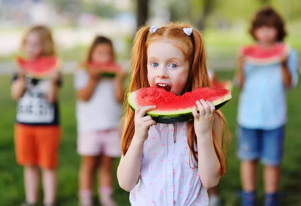 Рыжая маленькая девочка жадно ест сочный спелый арбуз на фоне своих друзей и Парк в солнечный летний день — стоковое фото