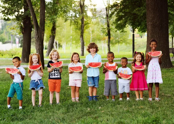 Группа счастливых детей дошкольного возраста разных национальностей держат в руках ломтики спелых арбузов и улыбаются на фоне парка в солнечный летний день. — стоковое фото