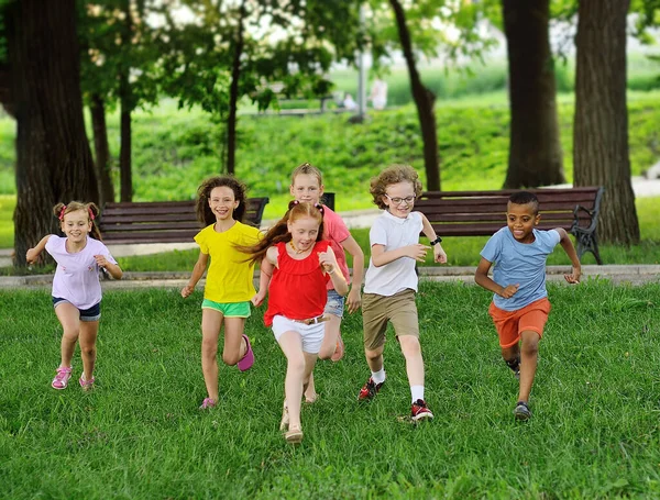 Farklı ırktan erkek ve kız çocuklarından oluşan bir grup yeşil çimlerde eğlence parkının arka planında koşuyor. Çocuk Günü, — Stok fotoğraf