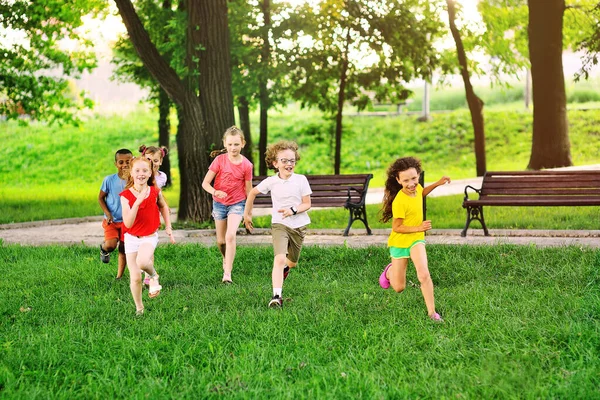 Bir grup anaokulu öğrencisi çimlerin üzerinde parkın arka planına ve yeşilliğe karşı koşuyor.. — Stok fotoğraf