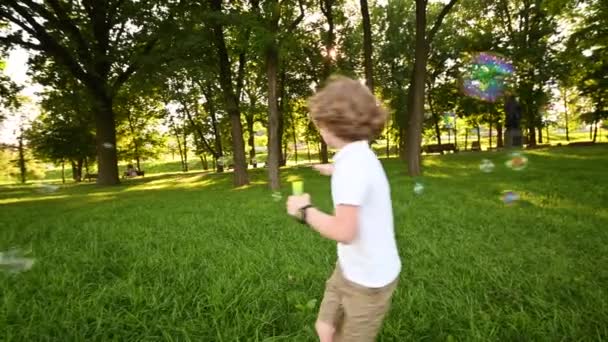Chlapeček v brýlích s kudrnatými vlasy foukání mýdlové bubliny a běh na pozadí zelené trávy a parku. — Stock video