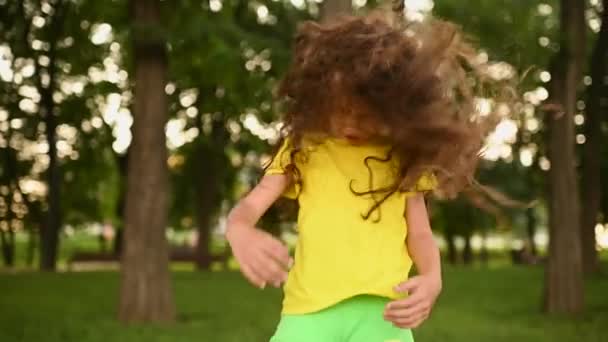 Aranyos kislány gyermek egy sárga póló göndör haj fordítja a fejét a háttérben a zöld és a park. — Stock videók