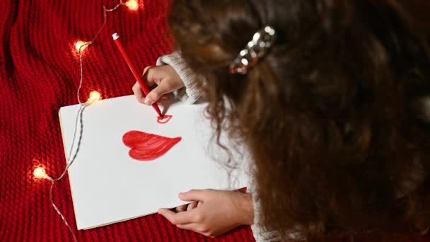 Een klein meisje met krullend haar tekent een hartje op een wit velletje papier voor Valentijnsdag liggend op een rode gebreide plaid. — Stockvideo