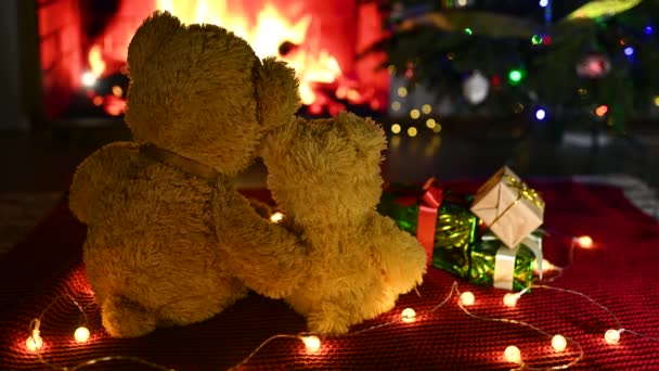 Dos bonitos osos de peluche abrazándose uno al otro sentados en un cuadros de punto rojo sobre el fondo de un árbol de Navidad, regalos y una chimenea ardiente — Vídeos de Stock