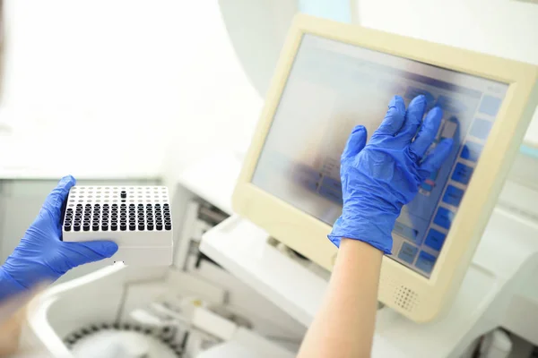 Un empleado de un laboratorio médico bacteriológico sostiene un recipiente con tubos de ensayo en sus manos contra el fondo del analizador. — Foto de Stock