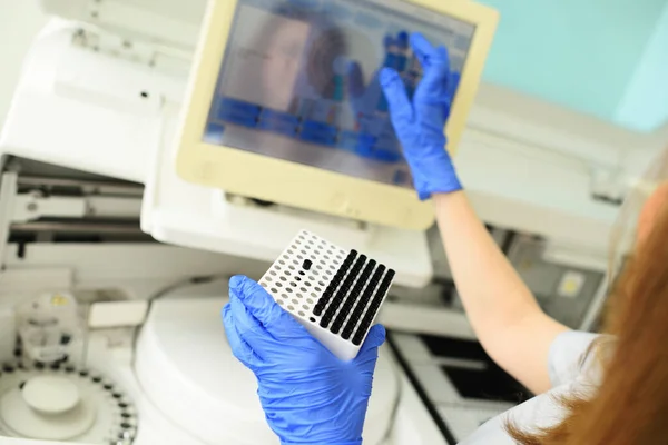 Un empleado de un laboratorio médico bacteriológico sostiene un recipiente con tubos de ensayo en sus manos contra el fondo del analizador. — Foto de Stock
