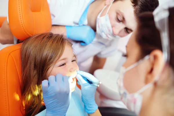 Fiatal fogorvosok, egy férfi és egy nő, megvizsgálja a fogak egy gyermek beteg - egy kis csinos lány, aki ül egy narancssárga fogászati szék. — Stock Fotó