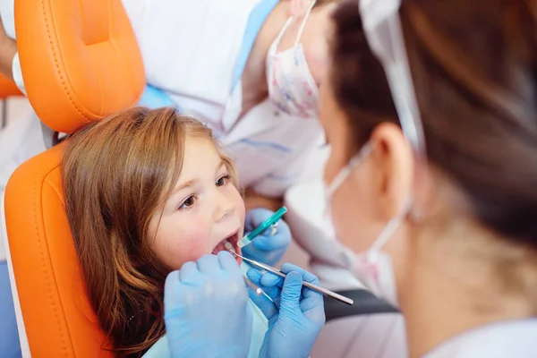 Dentistas jóvenes, un hombre y una mujer, examinan los dientes de un paciente infantil - una niña bonita que está sentada en una silla dental naranja. — Foto de Stock