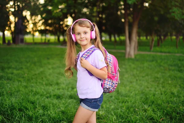 Uma menina bonito com uma mochila xadrez escola ouvir música em fones de ouvido rosa e sorrindo contra o fundo do parque e vegetação. — Fotografia de Stock